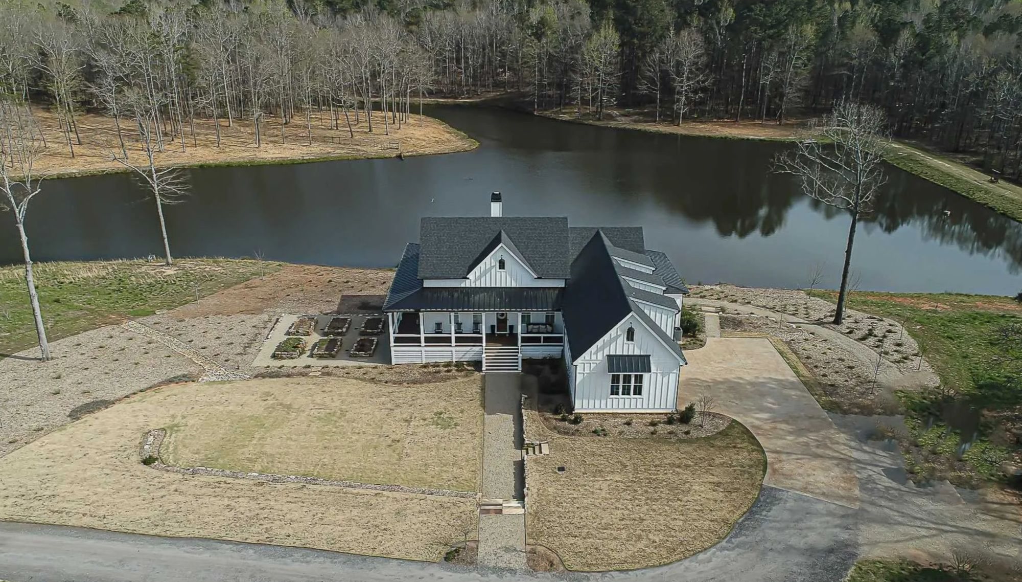 Aerial view of a white house with a wraparound porch on a lakeside property surrounded by bare winter trees.