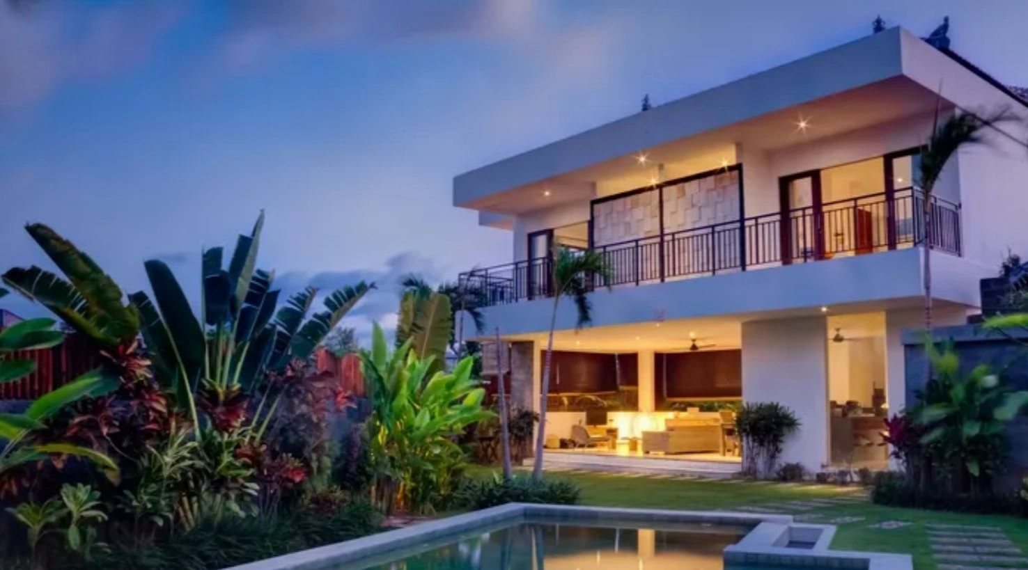 Twilight exterior of a modern home with a lit pool and tropical planting.